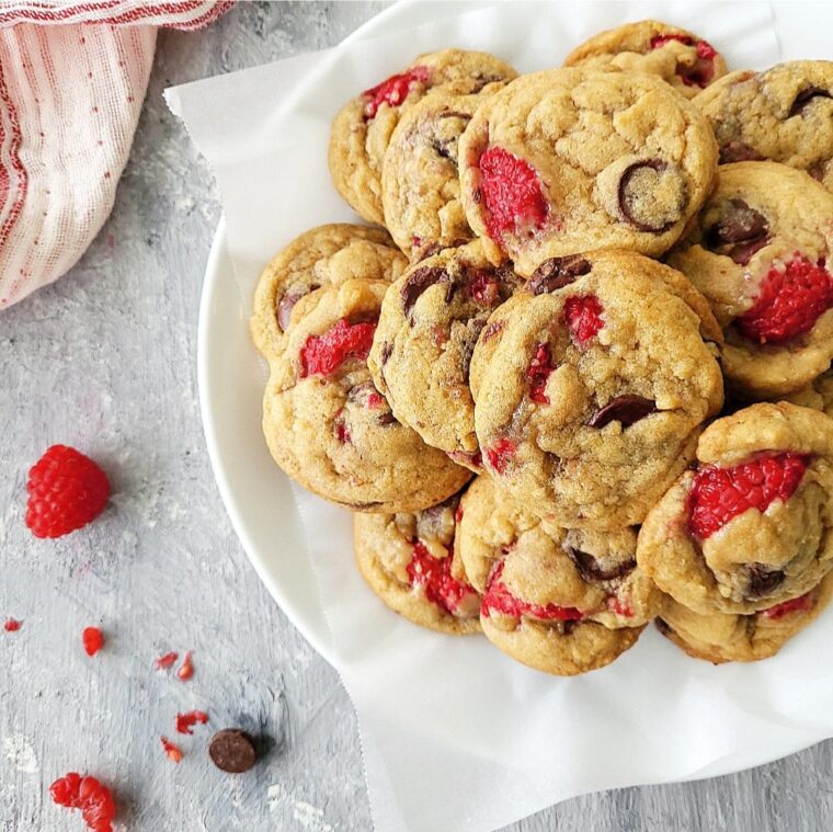 dark chocolate chip raspberry cookies stacked on a white plate top down photo styled with a pink striped linen, fresh raspberries and chocolate chips on a pale blue surface