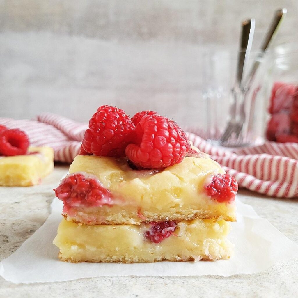 raspberry blondies side view of two blondie squares stacked on top of each other with a pile of fresh raspberries on top. background is gray scene is styled with a red and white striped tea towel, a glass with forks in it and a jar filled with fresh raspberries