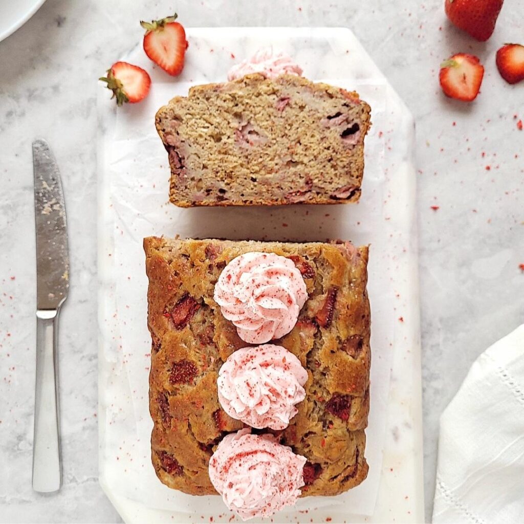 strawberry banana bread with strawberry buttercream rosettes top down view of loaf with one slice cut and facing upwards to showcase the crumb and strawberries. loaf is on a rectangular piece of white marble on a gray slab and styled with freshly cut strawberries and a stainless steel knife