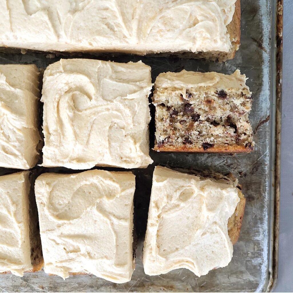 peanut butter cream cheese frosting on a banana chocolate chip sheet cake cake. top down view. cake is cut into large squares and one slice is turned on its side so you can see the moist inner crumb and chocolate chips