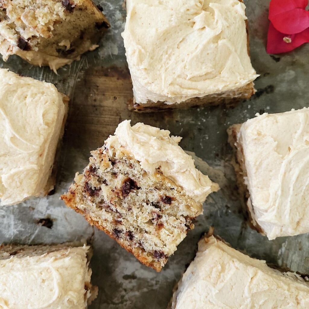 banana chocolate chip sheet cake with peanut butter cream cheese frosting. multiple slices cut into squares, top down photo with two slices on their sides so you can see the chocolate chip crumb detail. surface is a weathered tin baking sheet
