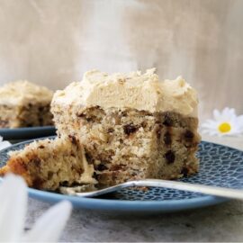 banana chocolate chip cake with peanut butter cream cheese frosting side view of 2 slices on blue plates one slice in foreground with a fork and one slice in back right corner. styled with daisies background is distressed brown and white