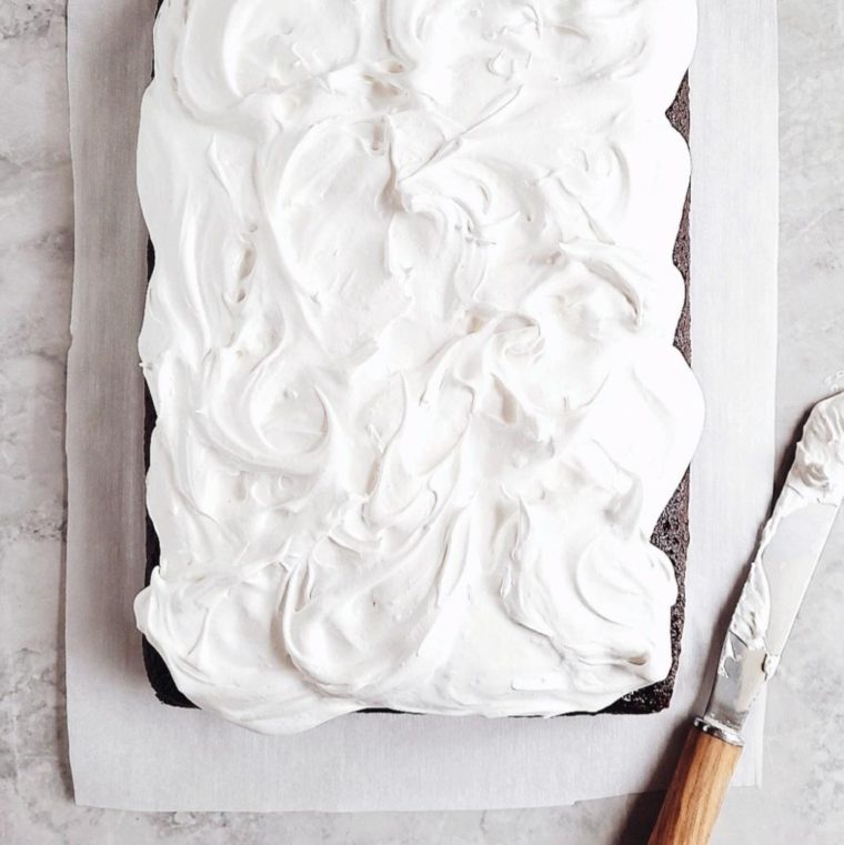 chocolate sheet cake with homemade marshmallow frosting top down view of uncut cake on a gray marble surface next to a wooden handle spatula with frosting all over it