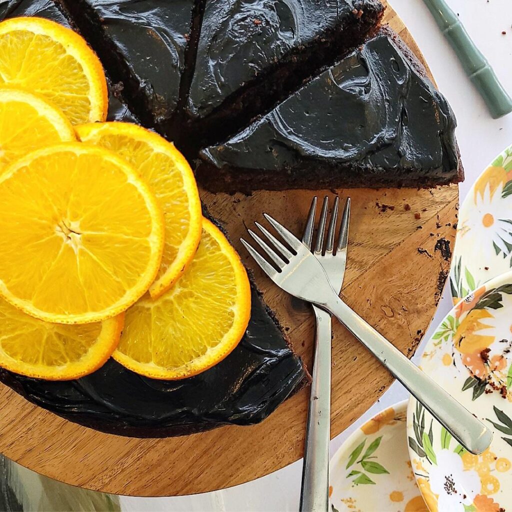 chocolate orange cake top down, close up view of cake on a wooden cake stand. cake is topped with black cocoa frosting. the left side of cake is uncut and adorned with a pile of sliced oranges. right side of cake is cut into 3 slices. there are 2 forks where a missing slice was. 