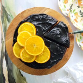 chocolate orange cake with black cocoa frosting top down view of cake on a wooden cake stand one slice has been cut and is being removed with a cake server. left side of cake has 6 orange slices as a garnish. top corner of photo is 3 white plates printed with orange flowers