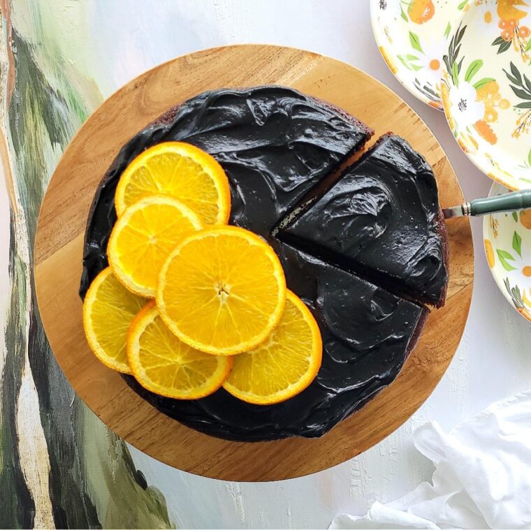 chocolate orange cake with black cocoa frosting top down view of cake on a wooden cake stand one slice has been cut and is being removed with a cake server. left side of cake has 6 orange slices as a garnish. top corner of photo is 3 white plates printed with orange flowers