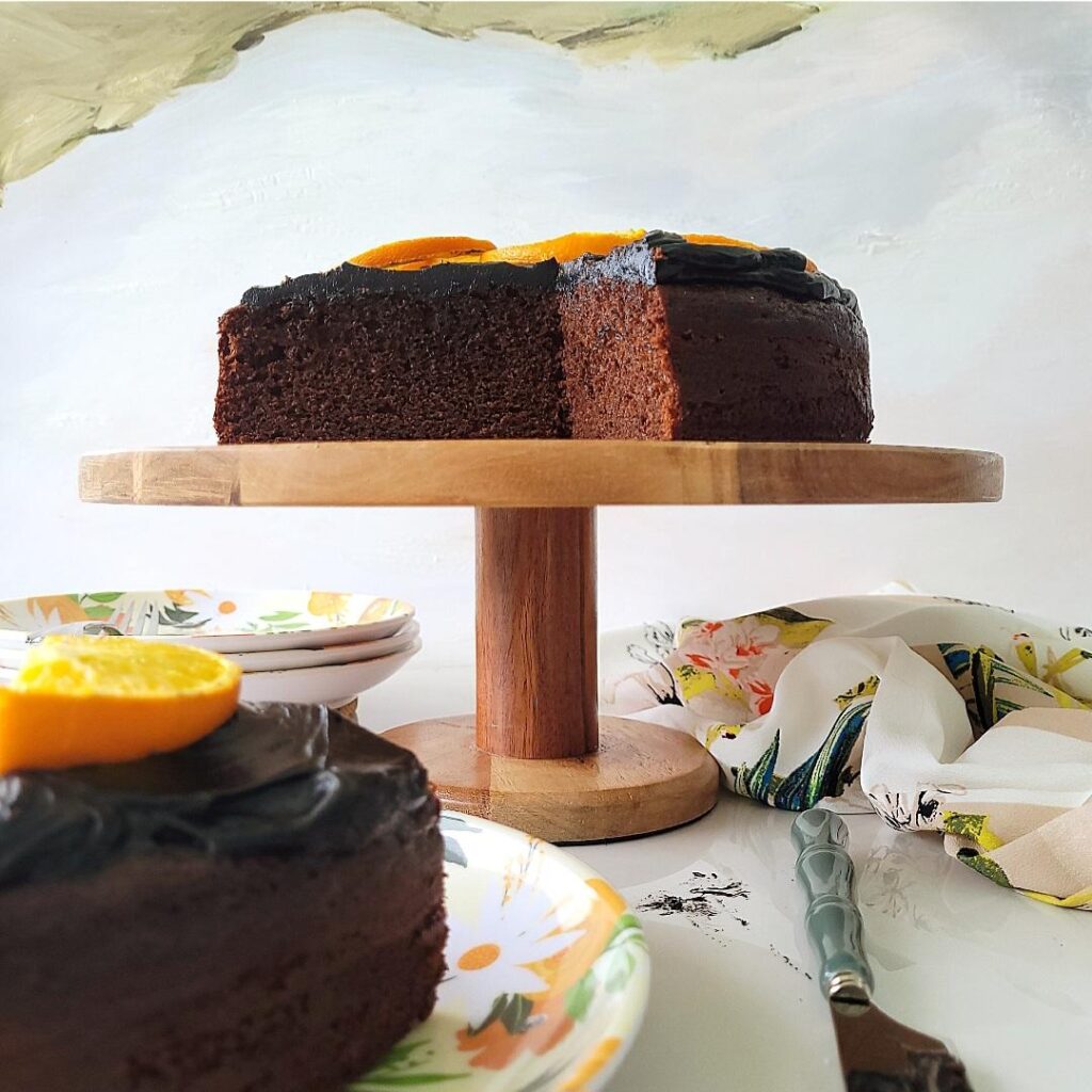 chocolate orange cake on a wooden cake stand. side view of cut cake so you can see the inner crumb. there is a cut slice on a flower patterned plate in the lower left foreground 