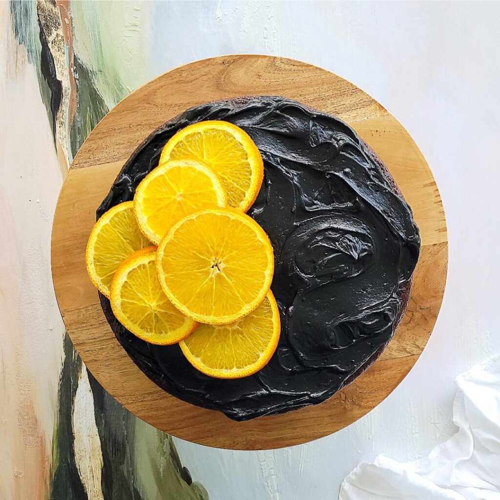 chocolate orange cake with black cocoa frosting and sliced oranges. top down view of uncut cake on a wooden cake stand. orange slices are on the left side of the cake only. 