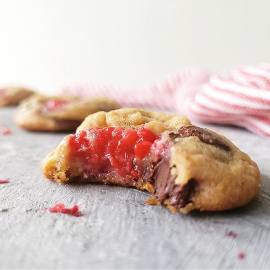 raspberry cookies with dark chocolate chips close up image of baked cookie with a bite missing so you can see the fruity, chocolaty gooey interior. background is white and styled with a pink striped linen and two cookies in the background