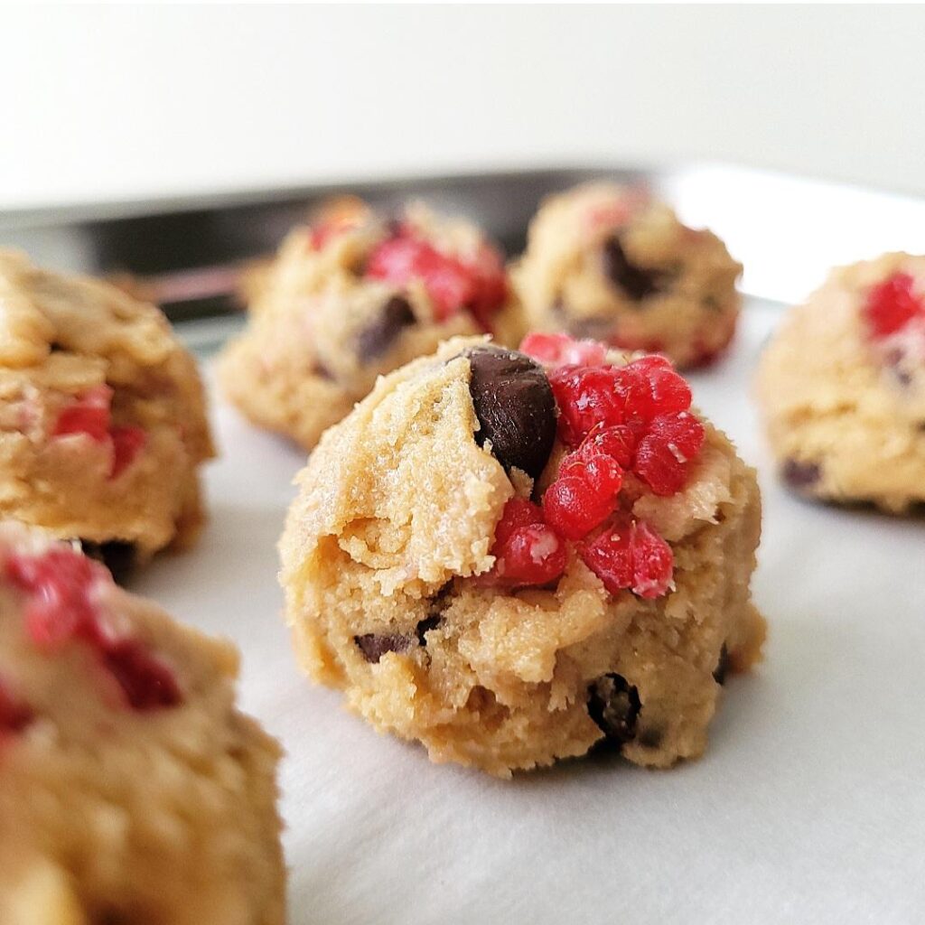 raspberry cookies with dark chocolate chips unbaked cookie dough balls on white parchment paper on a silver baking tin close up view of dough, chocolate chips and red raspberries