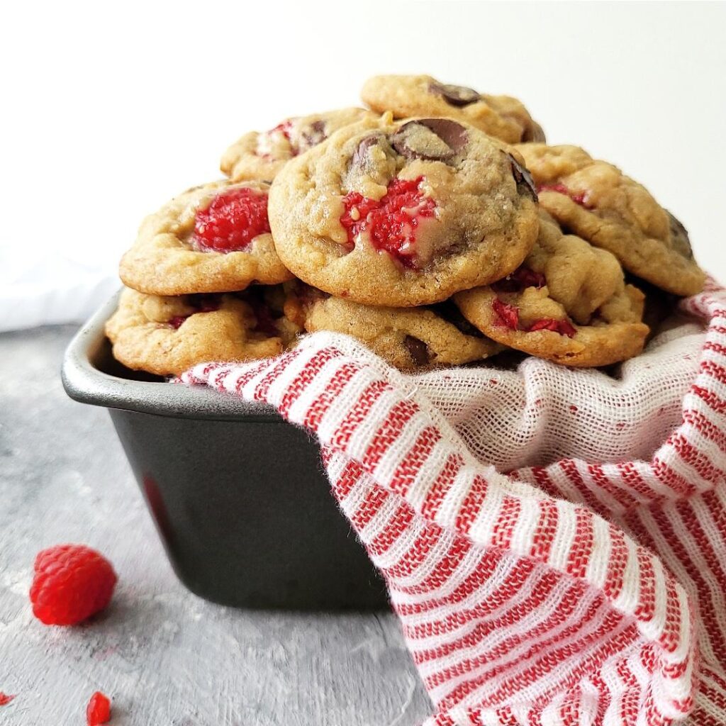 raspberry chocolate chip cookies piled high in a dark metal tin lined with a pink and white striped linen background is white and image is styled with a few fresh raspberries