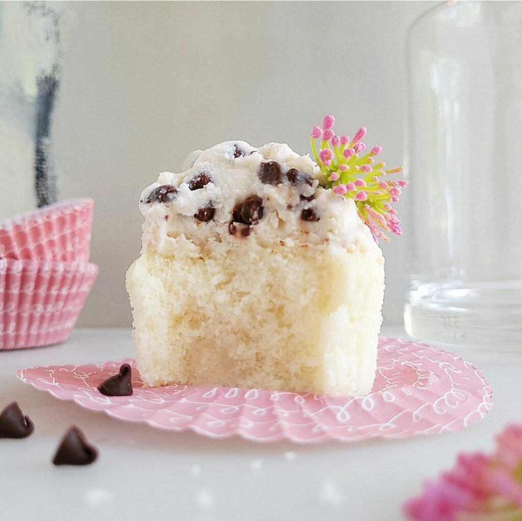 cannoli cupcake side view of vanilla cupcake topped with cannoli frosting with mini chocolate chips. cupcake has been bitten so you can see the fluffy crumb. frosting is garnished with a single pink flower. cupcake is sitting on a flattened pink cupcake wrapper