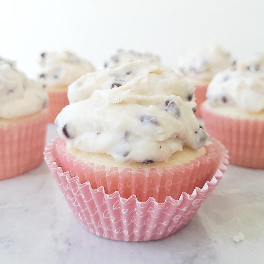 side view of 6 vanilla cupcakes topped with cannoli frosting. each cupcake has a pink cupcake liner. cannoli cream frosting is made of ricotta and mini chocolate chips