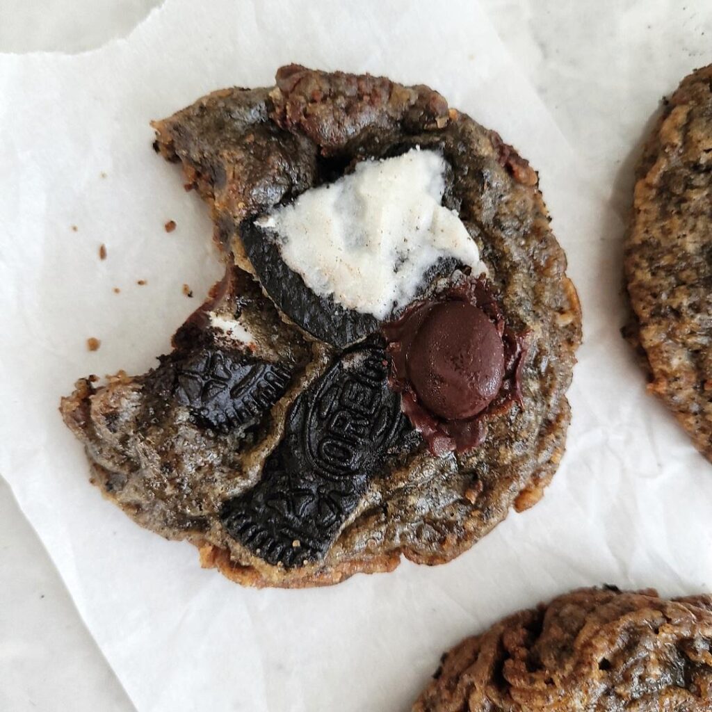 oreo chocolate chip cookie close up, top down view of one cookie with a bite missing. cookie is topped with big chunks of broken oreo cookies and chocolate chips