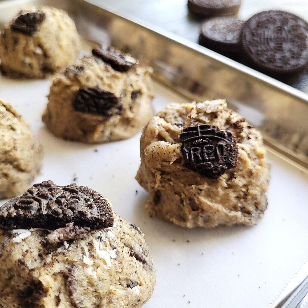 unbaked oreo chocolate chip cookies on a shiny metal baking sheet. close up of balled up cookie dough with broken pieces of oreo cookies on top