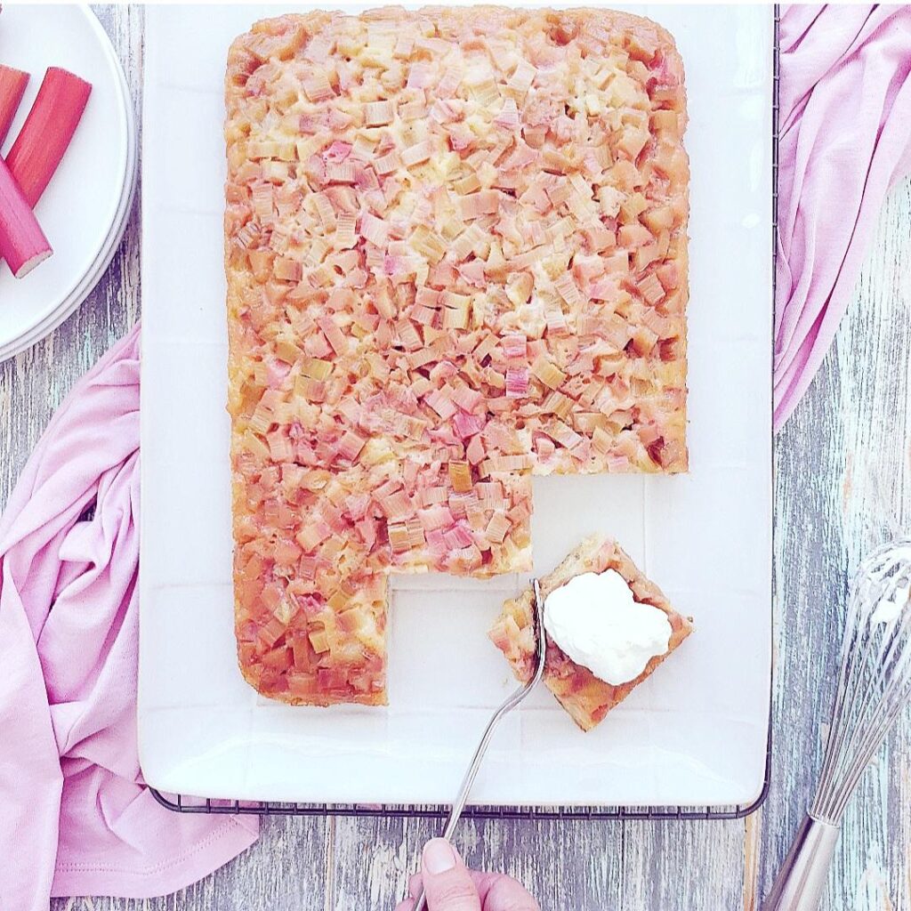 rhubarb upside down cake. top down view of a rectangular sheet cake topped with diced pink rhubarb. a square of cake has been cut from the bottom right corner of the white serving plate and is topped with vanilla ice cream. background is light blue distressed wood