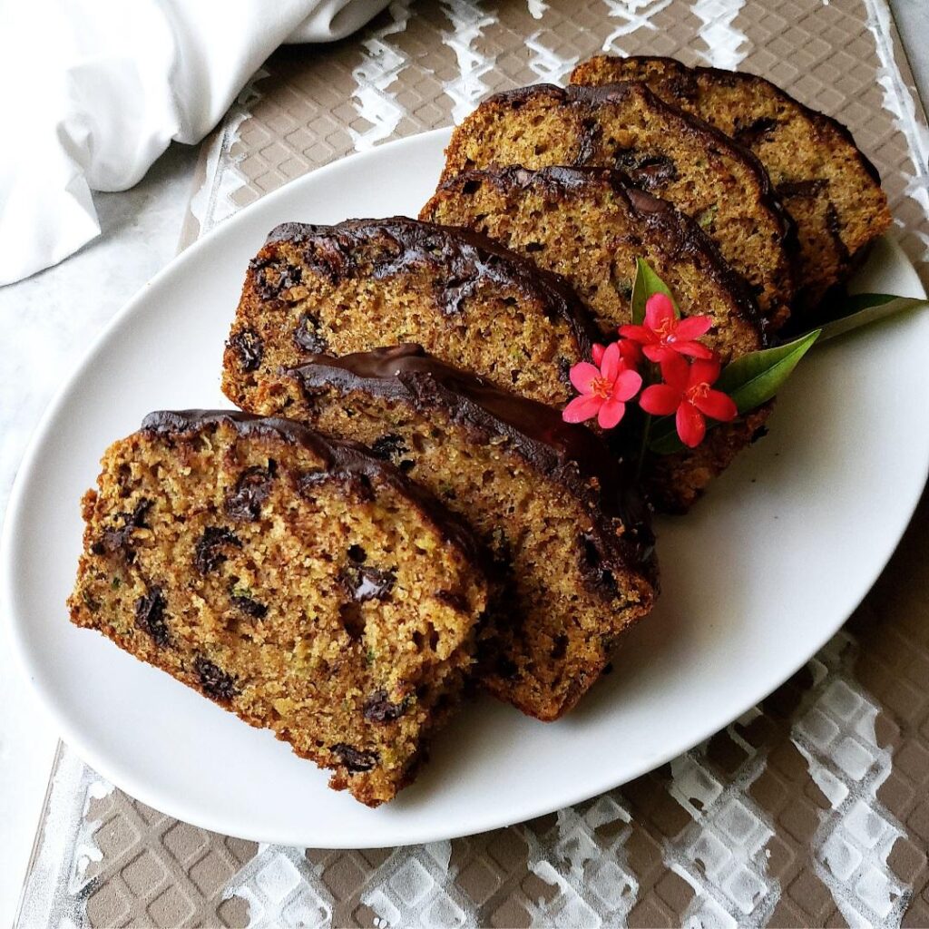 chocolate chip zucchini bread with chocolate ganache. loaf cut into slices and arranged on an oval white serving plate. bread is garnished with pink flowers. top down view.