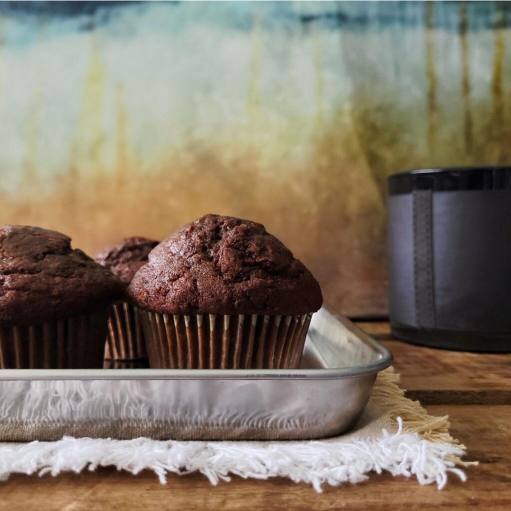 chocolate zucchini muffins on a shiny metal baking tray. side view background is abstract browns and golds. surface is distressed wood with a fringe linen. there is a black candle in the black right corner