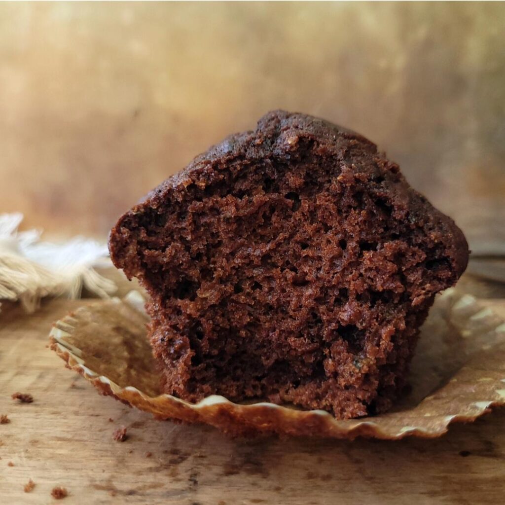 side view of a single chocolate zucchini muffin. muffin has been bitten so you can see the moist crumb inside. muffin is sitting on a brown muffin liner. background is abstract brown and tan. surface is distressed wood with muffin crumbs and there is a fringed linen in the back left corner.