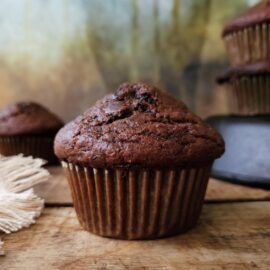 chocolate zucchini muffins. close up side view of bakery style chocolate zucchini muffin on a wooden surface. there is a stack of muffins in the back right and a single muffin in the back left of the frame. background is abstract painting that has brown and yellow hues.