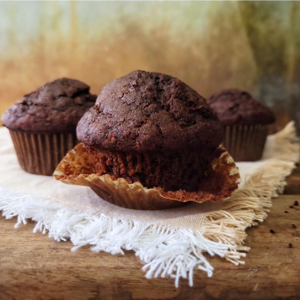 trio of chocolate zucchini muffins side view photo. front muffin has its liner pulled down so you can see the crumb. background is abstract brown and gold. surface is distressed wood with a fringed linen under the muffins.
