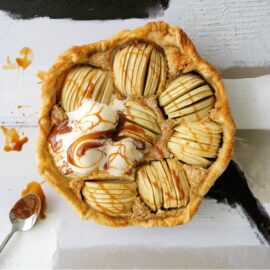 puff pastry apple tart with vanilla ice cream and salted caramel sauce. top down view of uncut tart styled with a spoon full of caramel on an abstract background that is white, black, gray and tan.