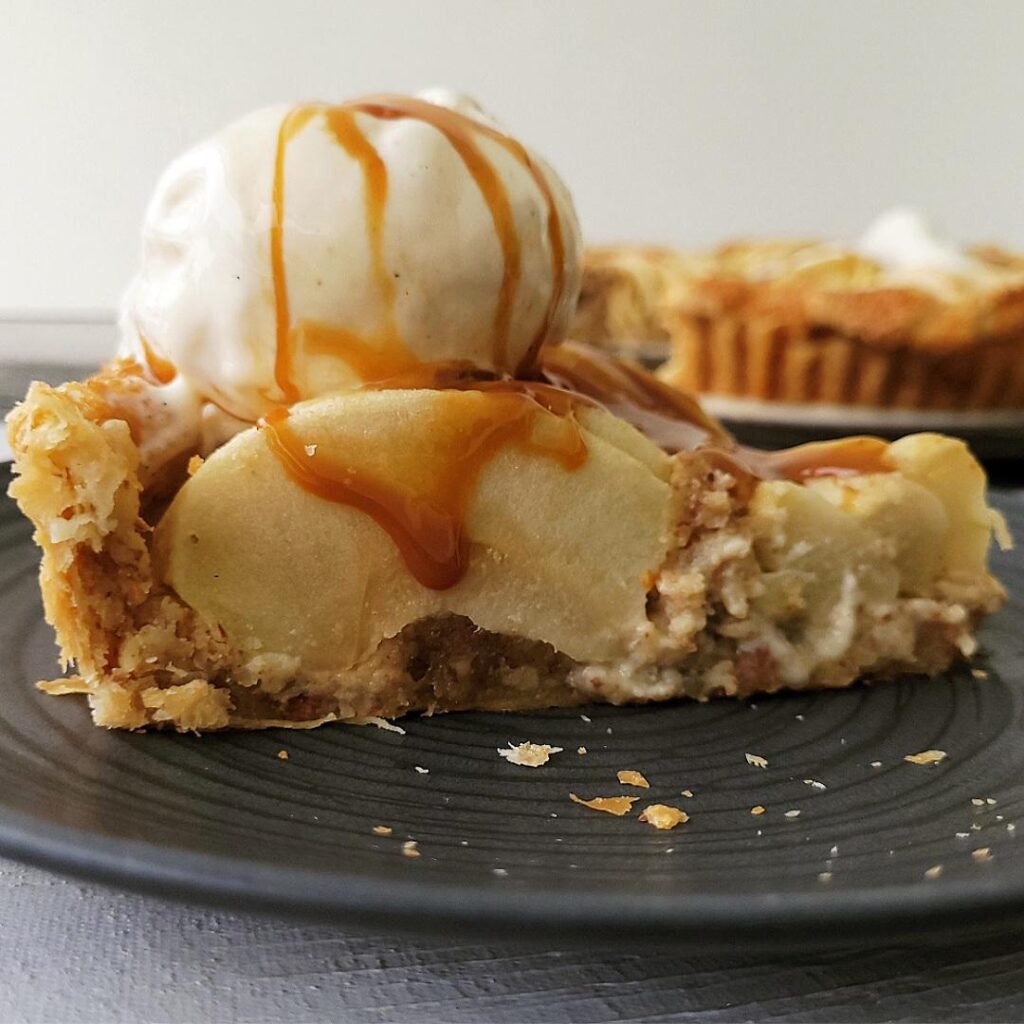 puff pastry apple tart. side view of one slice on a black plate. slice of apple tart is topped with a scoop of vanilla ice cream and dripping caramel sauce. 