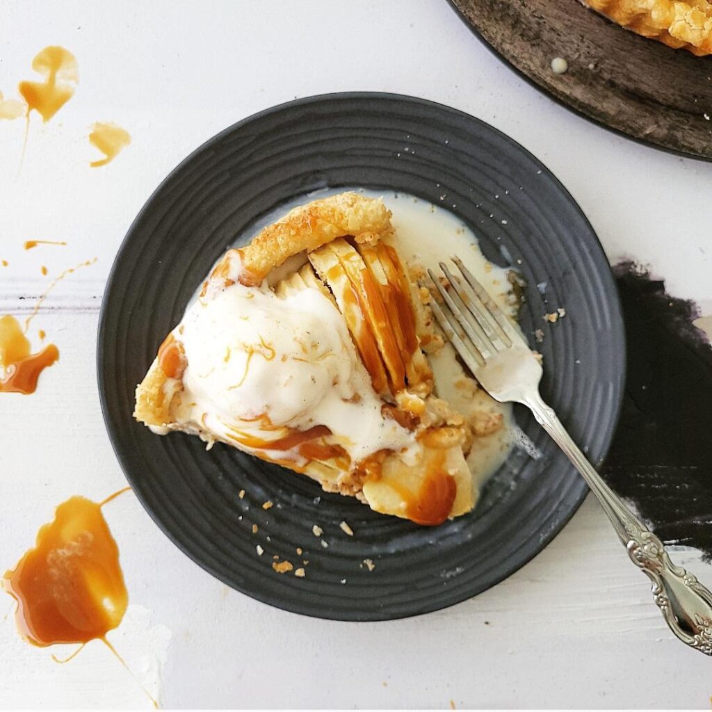 top down view of a slice of puff pastry apple tart on a black plate with a fork. tart is topped with a melty scoop of vanilla ice cream and caramel sauce. 