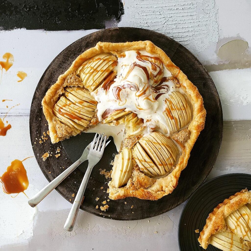 puff pastry apple tart. top down view of tart on a black wood charger with one slice cut away and there are two forks in the void space. tart is topped with melty vanilla ice cream and swirls of caramel sauce. there is a black plate in the lower right corner with a slice of apple tart on it.