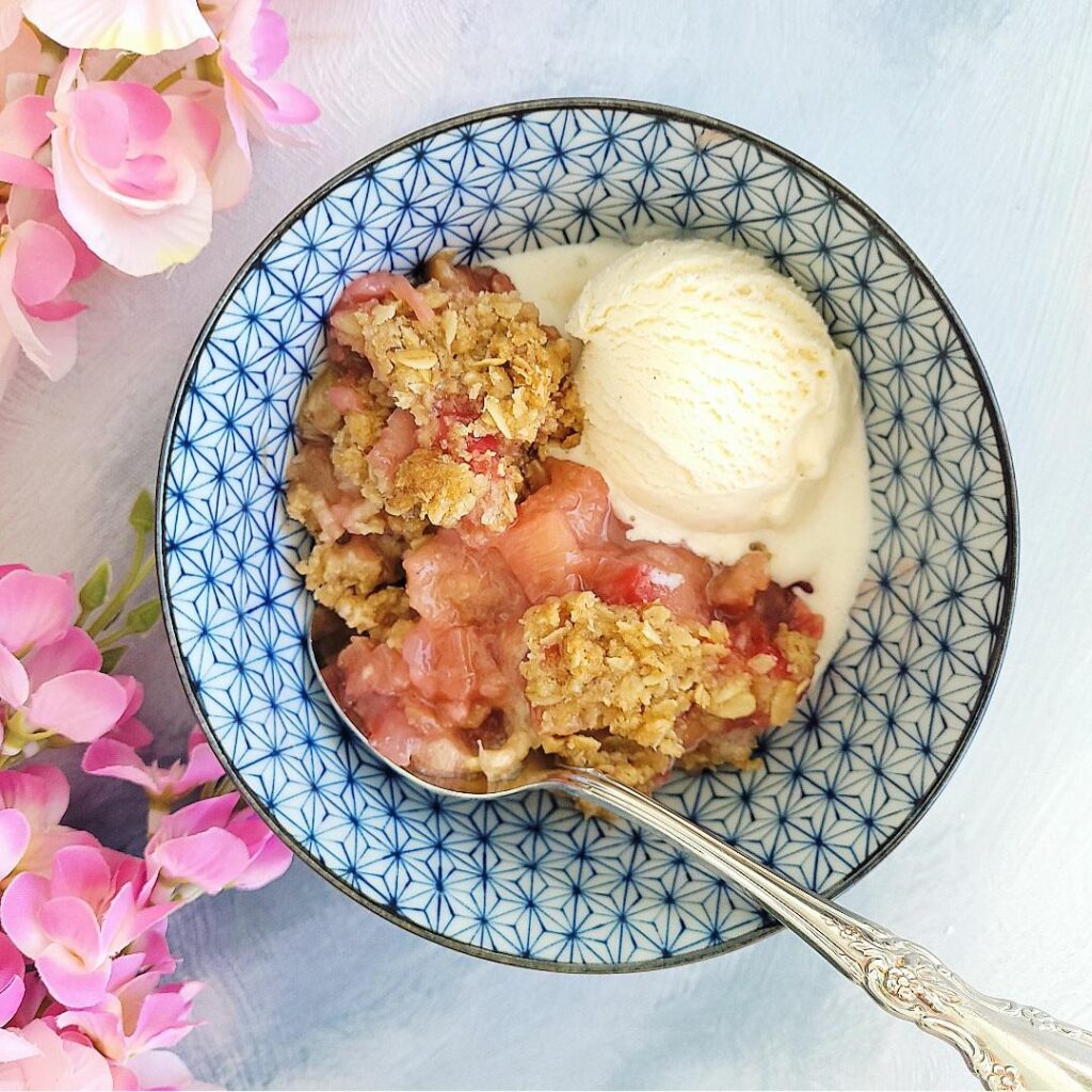 rhubarb crisp in a pretty blue bowl with a scoop on vanilla ice cream and a spoon. top down view. bowl is next to pretty pink flowers
