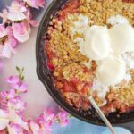 cast iron skillet rhubarb crisp top down view of crisp with three melting scoops of vanilla ice cream. skillet is surrounded by pink flowers