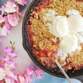 cast iron skillet rhubarb crisp top down view of crisp with three melting scoops of vanilla ice cream. skillet is surrounded by pink flowers