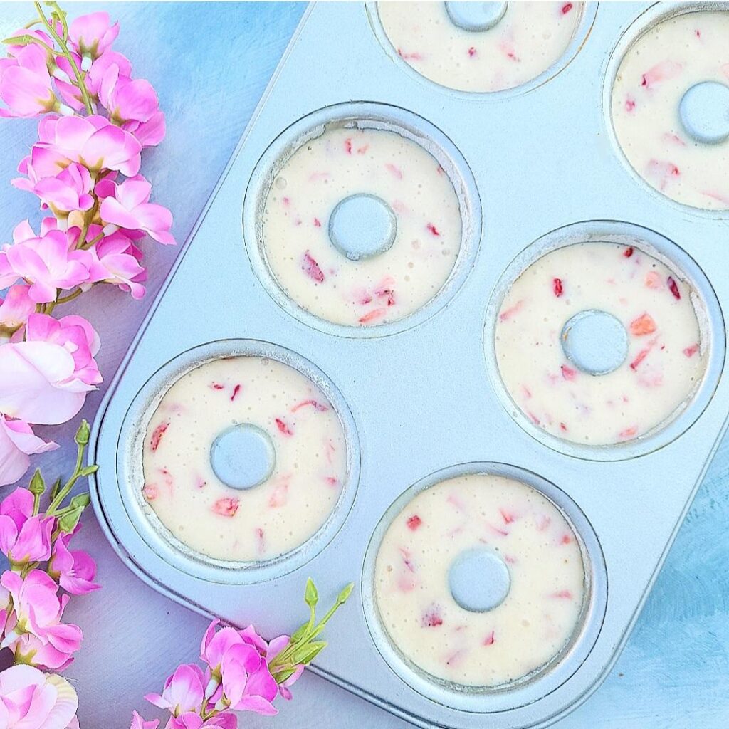 strawberry donuts. top down photo of batter in a metal donut pan. background is blue and there are pink flowers on the left of the frame.
