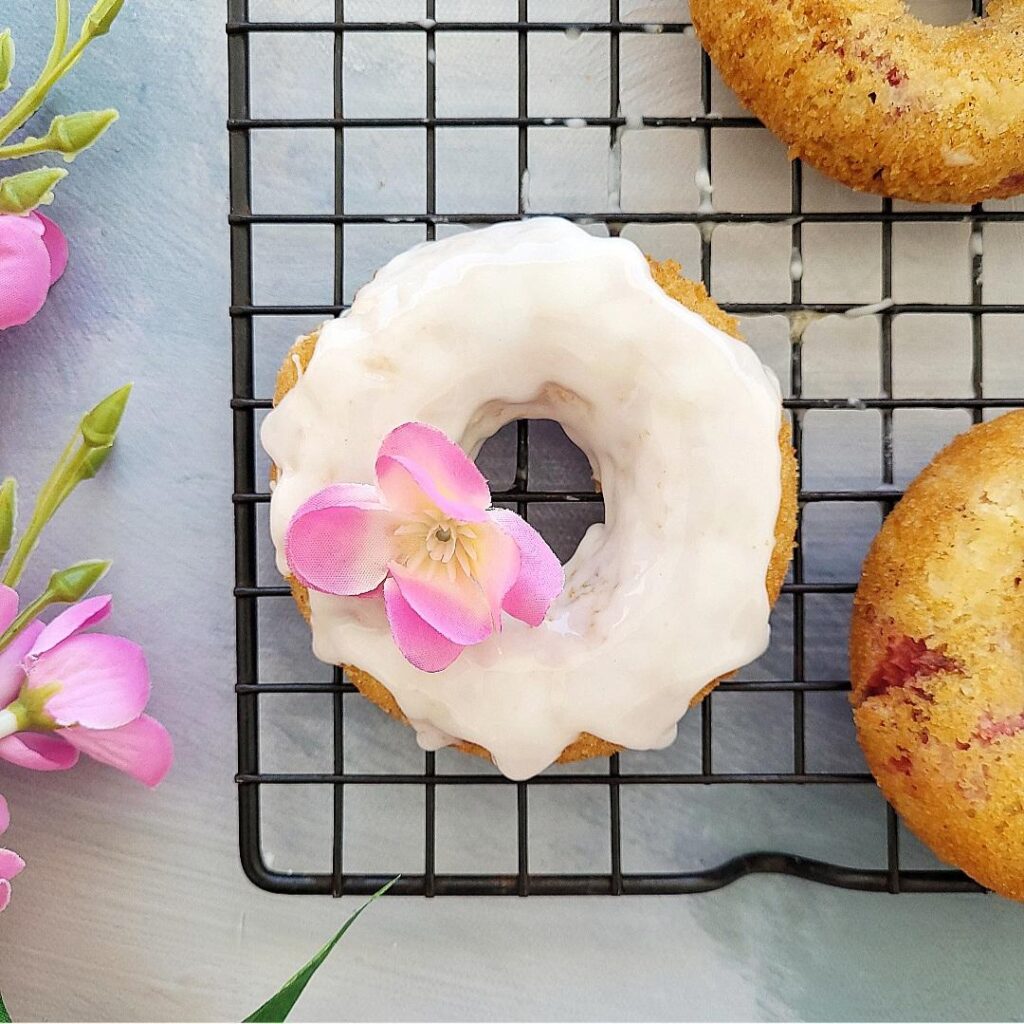 baked strawberry donuts. top down, close up view of one donut with white glaze and a pink flower on a black baking rack.  two other donuts are partially visible on the right. there are pink flowers on the left.