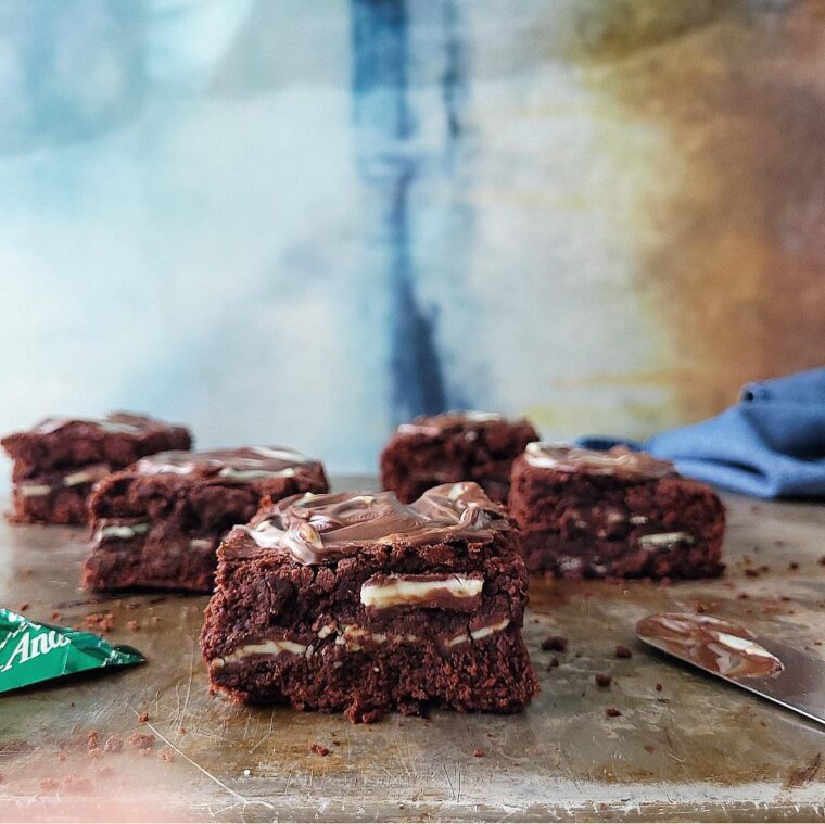 andes mint brownies. side view of 5 cut brownies on a metal baking pan. layers of brownie and mint candies are visible. background is abstract blue white and brown.