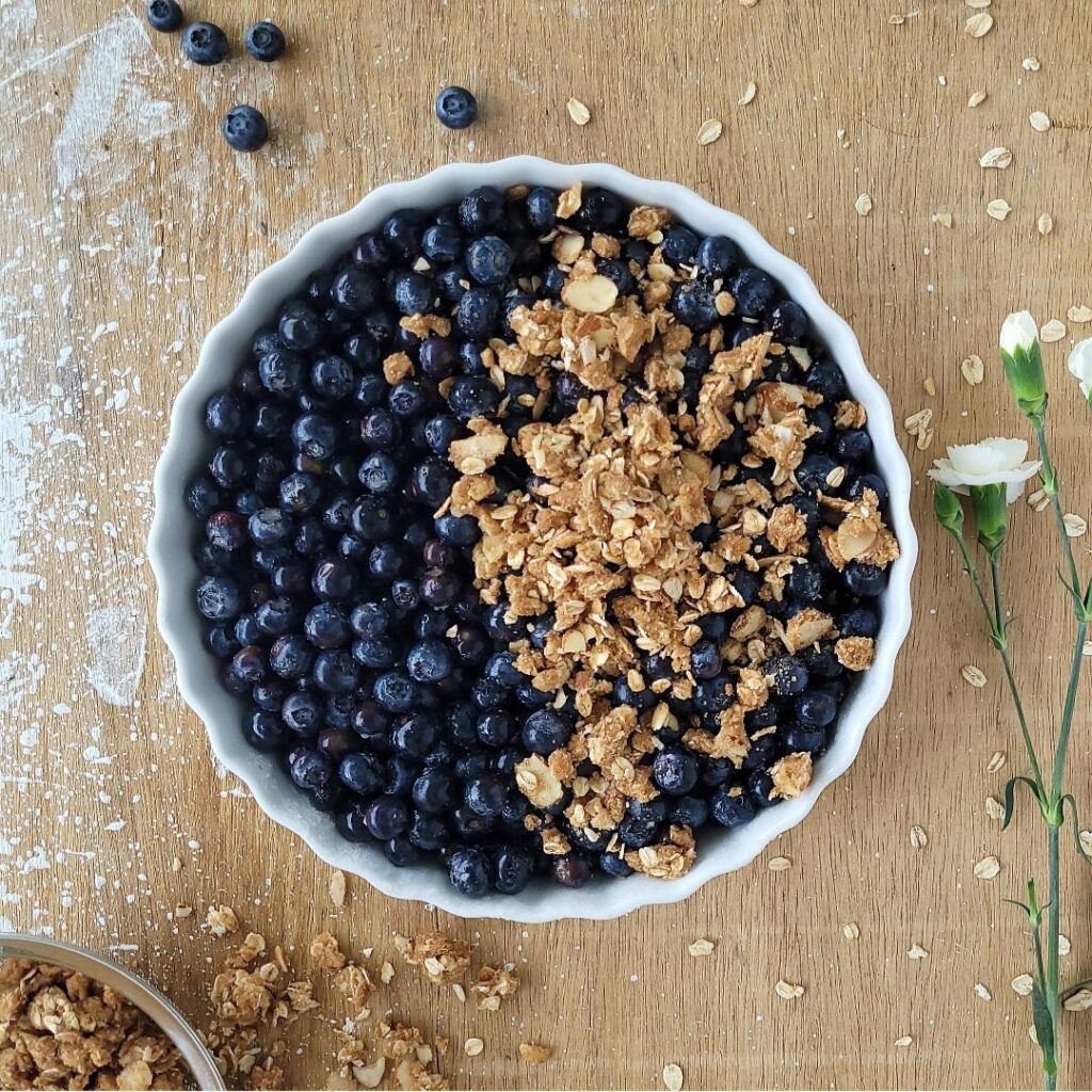 blueberry almond crisp. top down view of tart before baking. background is wood with white speckles of paint and styled with fresh blueberries and tiny white carnations. center of frame is close up of blueberries in a white ceramic tart plate. the right half of the tart has been sprinkled with almond oat topping.