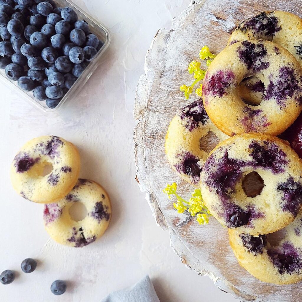 baked blueberry donuts top down view. soft fluffy donuts spotted with blueberries baked in. donuts are piled on a round wooden charger with distressed white paint and styled with tiny yellow flowers. there is a container of blueberries in the upper left corner.