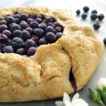 blueberry galette with fresh blueberries and a turbinado sugar crust on this rustic pie. three quarter angle, close up view of the galette. image is styled with fresh blueberries and white flowers.