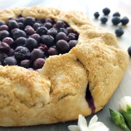 blueberry galette with fresh blueberries and a turbinado sugar crust on this rustic pie. three quarter angle, close up view of the galette. image is styled with fresh blueberries and white flowers.