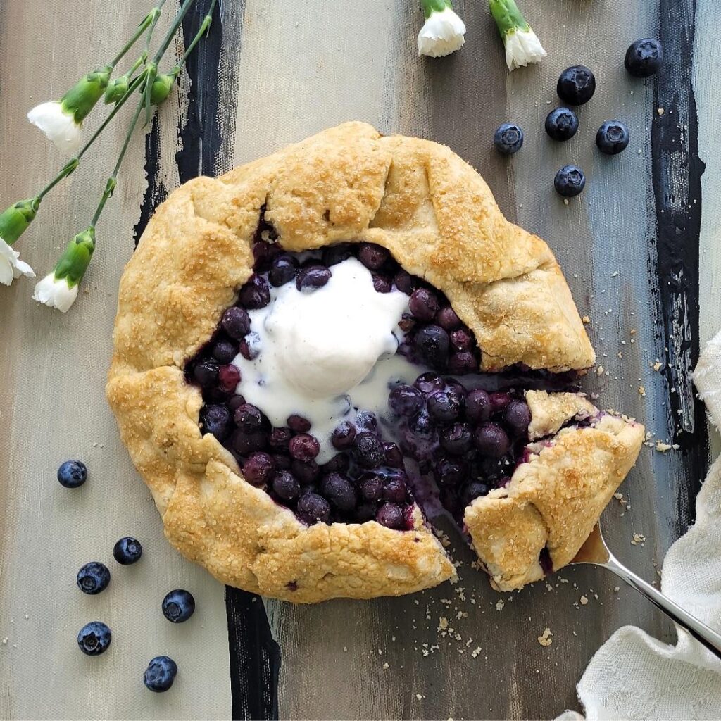 blueberry galette top down view of galette with a single slice cut in the lower left. slice is resting on a silver pie server. galette has a melting scoop of vanilla ice cream on top. background is abstract and styled with fresh blueberries, a cream linen and tiny white carnations. 