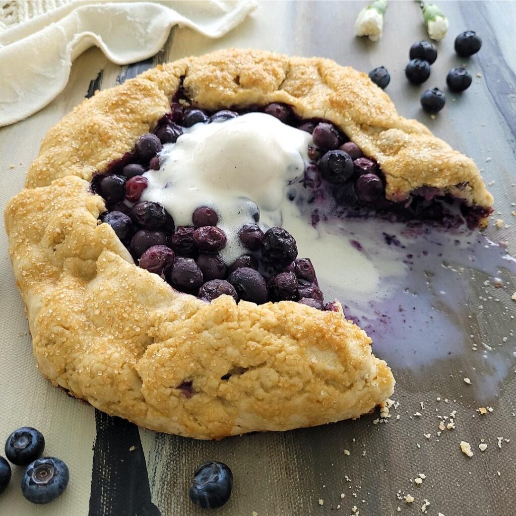 blueberry galette with a melty scoop of vanilla ice cream on top. one slice has been cut and removed from galette and the ice cream is melting in the void space. background is styled with fresh blueberries a cream linen and tiny white carnations. 