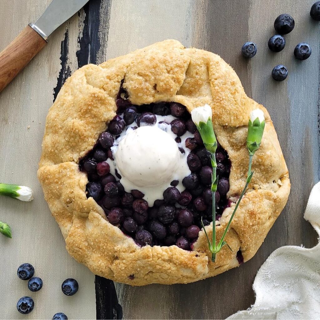 blueberry galette with a scoop of vanilla ice cream. top down view of uncut galette garnished with two small white carnations. background is blue, gray and black abstract and is styled with fresh blueberries, a wooden-handle spatula and a cream linen.
