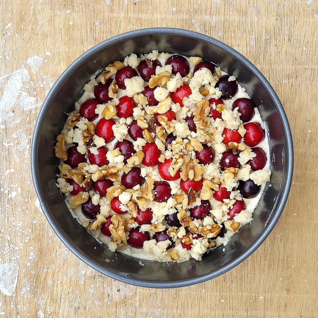 cherry buckle cake. top down view of batter in a metal springform pan with fresh cherries and the crumbly butter walnut topping before baking. background is a wooden surface with white paint splattered on it.