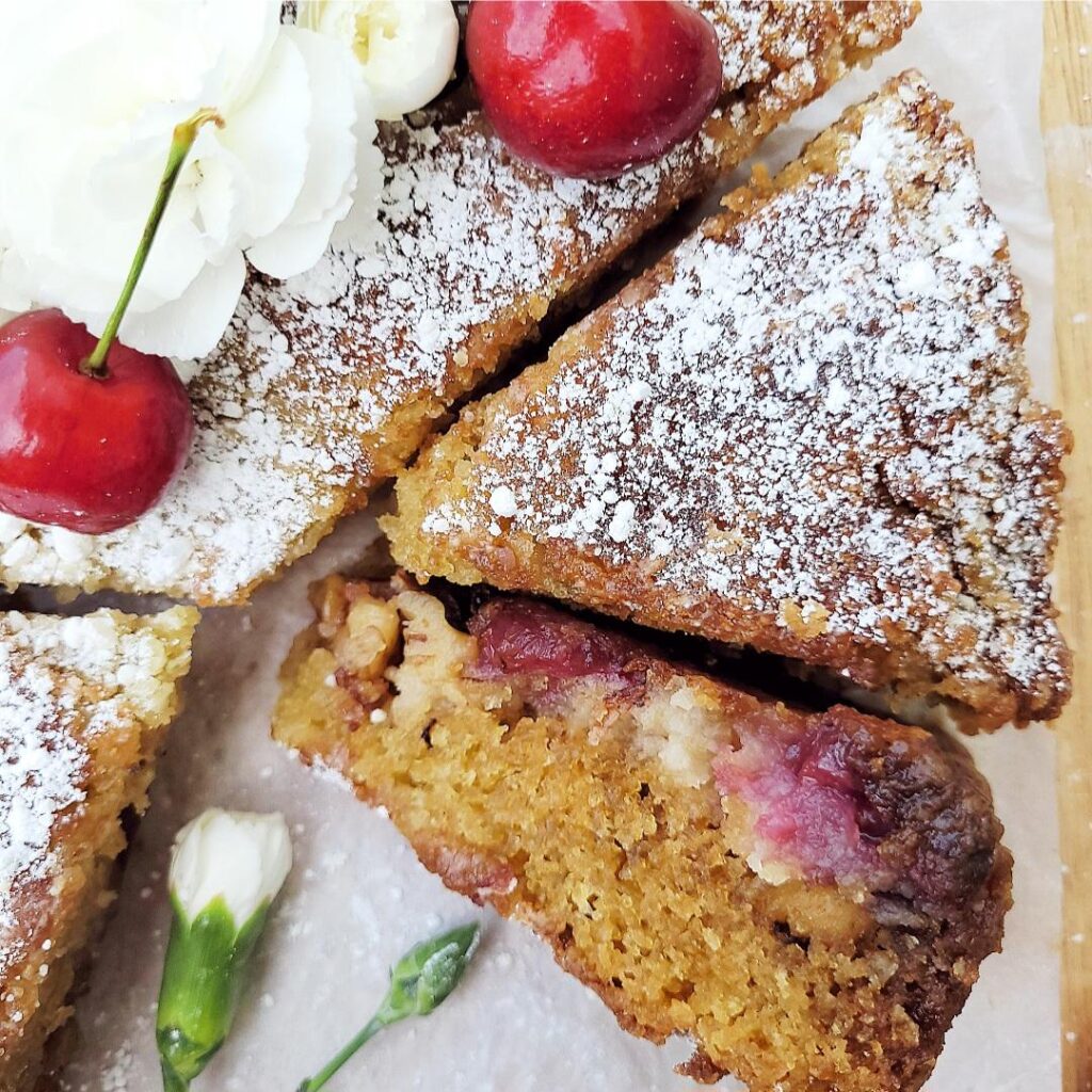 cherry buckle cake. zoomed in, top down view of cake two slices are cut and one is flipped on its side so you can see the crumb, walnuts and cherries baked in. cake is dusted with powdered sugar and garnished with stemmed cherries and white carnations. 