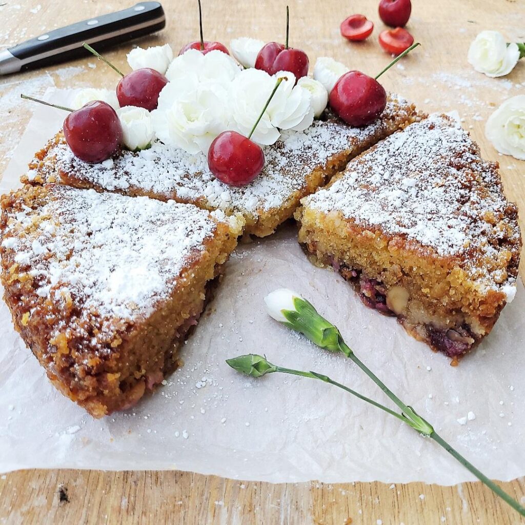 cherry buckle cake. 3/4 view of cake two slices are cut and a 3rd slice is missing. cake is topped with white powdered sugar, stemmed cherries and tiny white carnations. angle is so you can see the interior crumb, walnuts and cherries baked in the cake. background is wooden and styled with more white flowers, cherries and a black knife handle. 