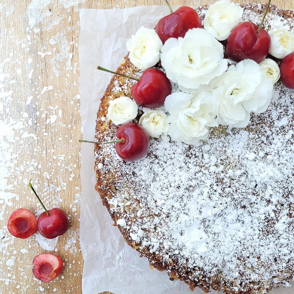 cherry buckle cake. top down view of a round, single layer cake topped with powdered sugar, tiny white carnations and fresh stemmed cherries. 