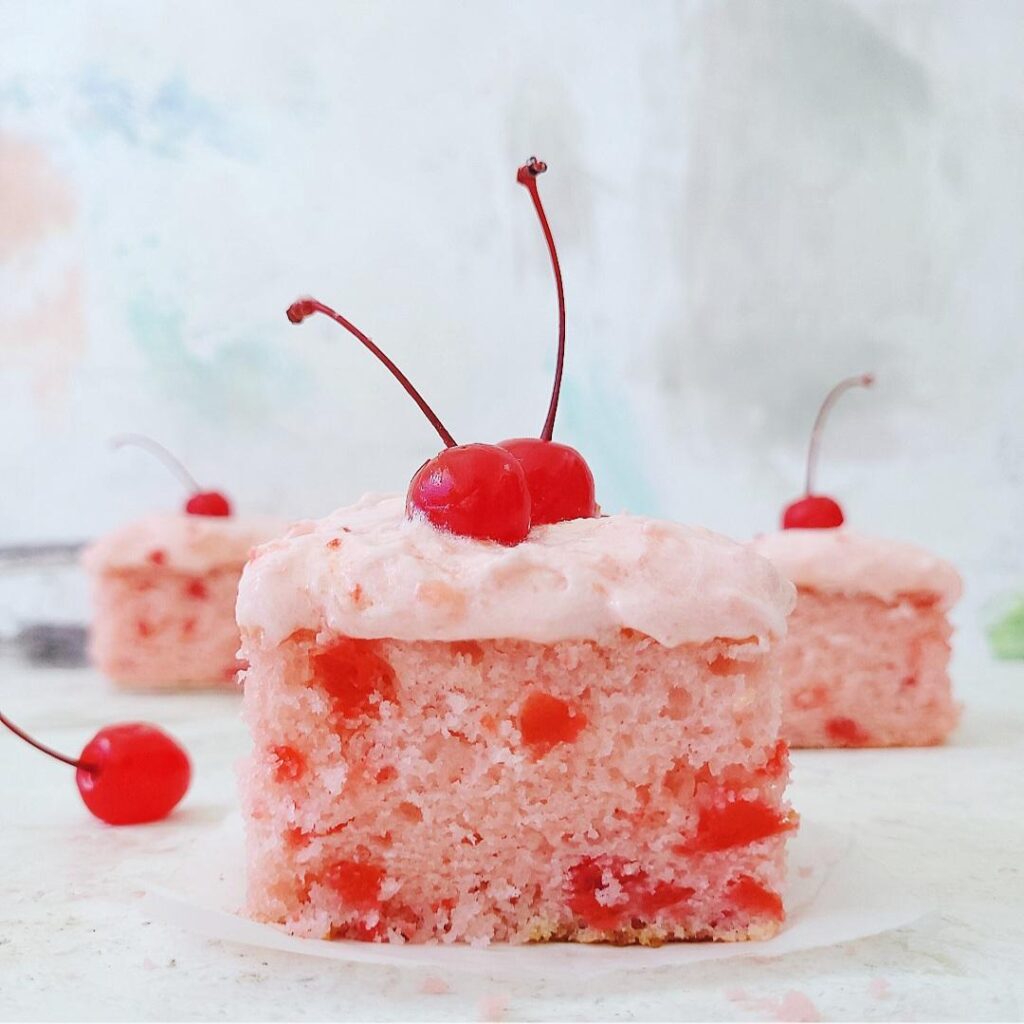 homemade cherry chip sheet cake with cherry frosting side view of three square slices so you can see the pink crumb and flecks of chopped maraschino cherries baked inside. each slice is topped with a stemmed maraschino cherry.