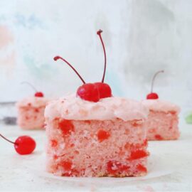 cherry chip cake. side view of three slices of sheet cake one in front and two in back. zoomed in view of pink crumb flecked with cherry bits. each slice is topped with cherry frosting and a stemmed maraschino cherry