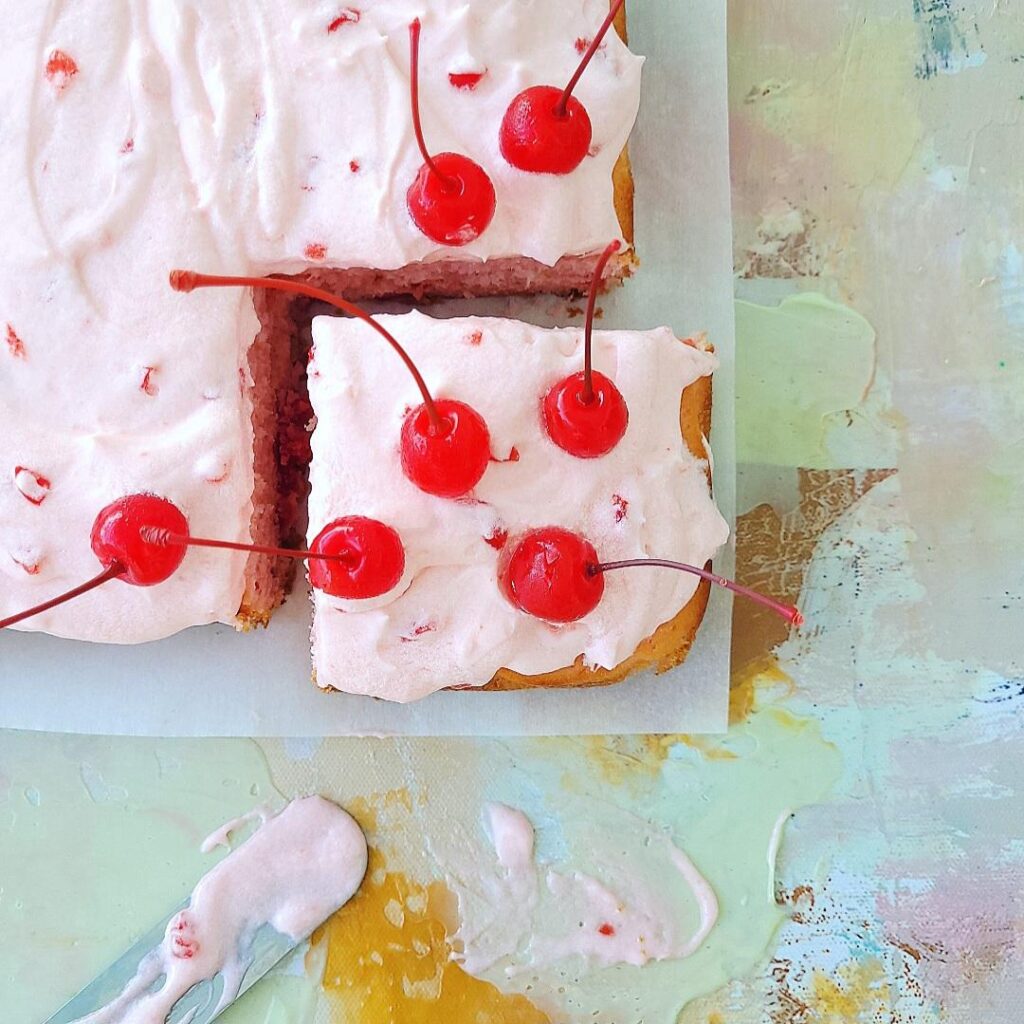 maraschino cherry chip cake. top down view of sheet cake with pink cherry frosting and maraschino cherries on top. close of image of a cut corner piece of cake. background is abstract art in pinks and whites.