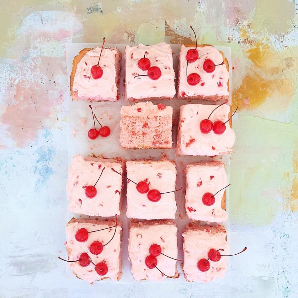cherry chip cake with cherry frosting. top down view of sheet cake cut into 12 slices. one slice is missing and replaced with two maraschino cherries. one slice is flipped on it's side so you can see the pink crumb flecked with cherry chip bits